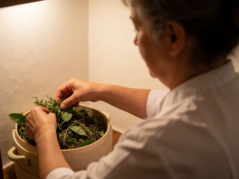 Des mains déposent des herbes fraîches dans un récipient en céramique sur une table, éclairage chaleureux et ambiance relaxante.