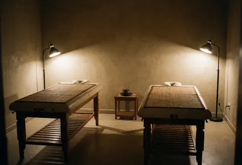 Salle de massage ayurvédique avec deux tables en bois, lampes sur pied et table d'appoint avec encens, ambiance relaxante et professionnelle.