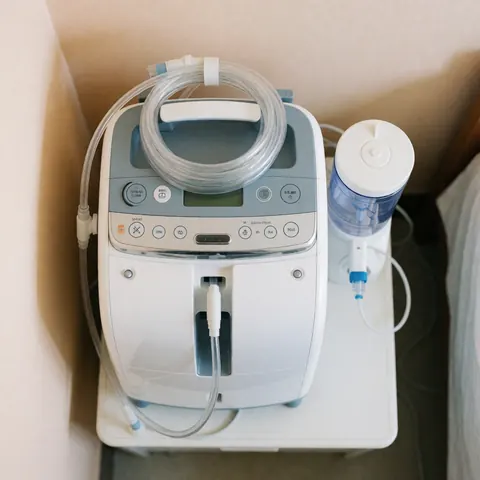 Un concentrateur d'oxygène médical blanc posé sur une surface claire avec son tuyau et un humidificateur, évoquant le soin à domicile.