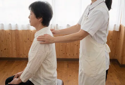 Un thérapeute en uniforme blanc réalise un massage shiatsu sur les épaules d'un client assis, dans un cadre lumineux aux murs en bois clair, évoquant relaxation et professionnalisme.