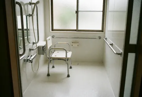 Intérieur d'une salle de bain sécurisée et accessible équipée d'une chaise de douche, de barres d'appui, d'un sol antidérapant et d'une fenêtre laissant entrer la lumière naturelle.