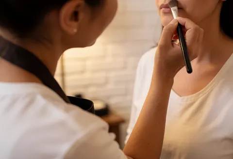 Maquilleuse professionnelle applique du maquillage sur une cliente à l'aide d'un pinceau dans un cadre chaleureux aux murs de briques blanches.