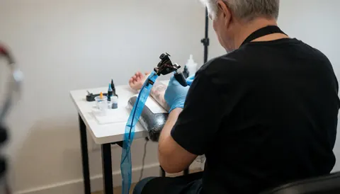 Un tatoueur professionnel avec des gants bleus applique de l'encre sur un bras posé sur une table blanche, entouré d'équipement stérile et d'un fond épuré.