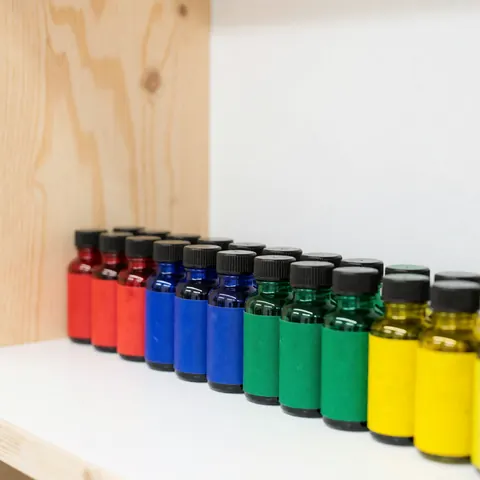 Des bouteilles d'encre végétale aux couleurs vibrantes sont alignées sur une étagère blanche, posée devant un fond en bois clair.