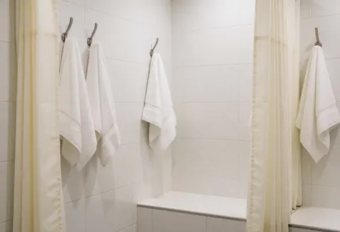 Intérieur d'une salle de douche équipée de rideaux et de serviettes blanches suspendues, murs carrelés blancs et banc en céramique pour un espace propre.