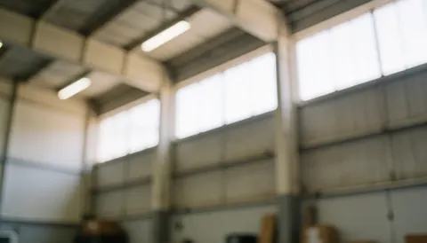 Arrière-plan flou d'un entrepôt industriel avec des poutres métalliques, des fenêtres hautes et une lumière naturelle douce, idéal pour du texte commercial.