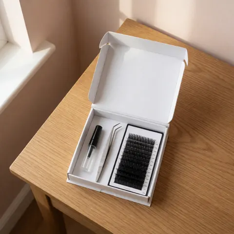 Kit de faux cils dans une boîte ouverte avec pince et tube de colle sur table en bois clair éclairée par la lumière naturelle.