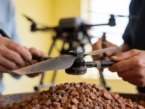 Un professionnel examine une hélice de drone poussiéreuse posée sur un tas de minerai rouge, avec le drone et des murs jaunes en arrière-plan.