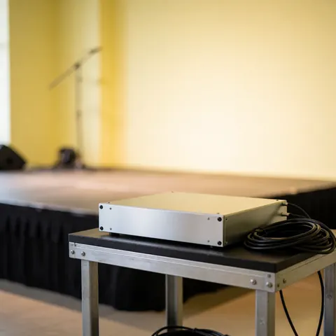 Enceinte sonore argentée posée sur une table métallique avec un câble enroulé, devant des murs jaunes et un arrière-plan flou.