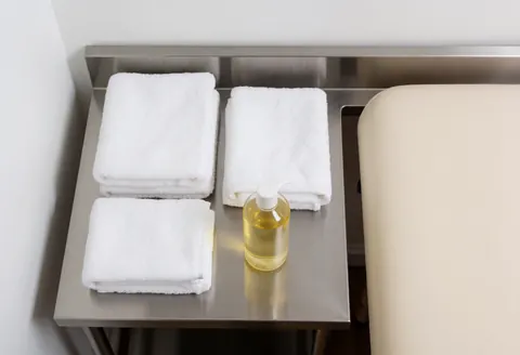 Table en acier inoxydable avec des serviettes blanches pliées et une bouteille d'huile de massage, près du bord d'une table de massage beige dans un environnement propre et professionnel.