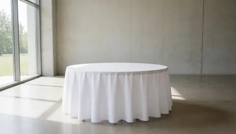 Une table ronde recouverte d'une nappe blanche se trouve dans une salle de événements aux murs en béton et à la grande fenêtre laissant entrer la lumière naturelle.