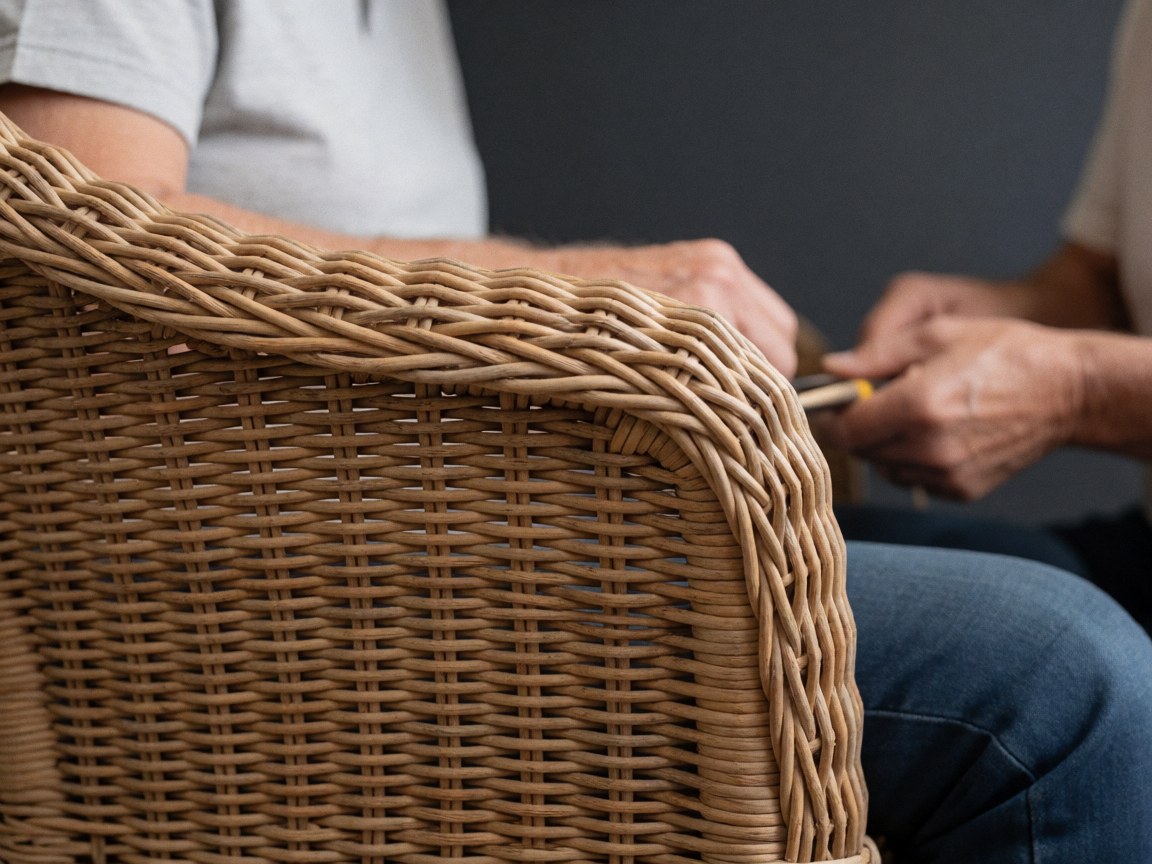 Gros plan sur des mains utilisant un outil pour réparer ou tisser une chaise en osier naturel, devant un mur gris anthracite.