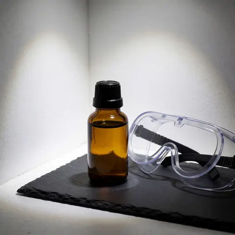 Flacon en verre ambré avec bouchon noir et lunettes de protection transparentes sur surface noire, éclairage dramatique dans un coin blanc.