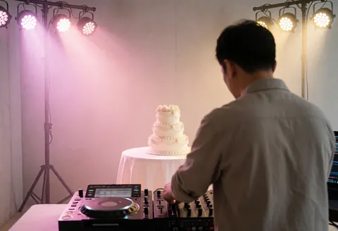 Un dj professionnel ajuste son matériel audio lors d'une fête, avec un gâteau de mariage en arrière-plan éclairé par des lumières roses et dorées.