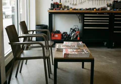 Espace d'attente dans un atelier de vélos avec deux chaises modernes et une table posant des magazines. Arrière-plan montrant un établi et des outils. Éclairage naturel par la fenêtre.