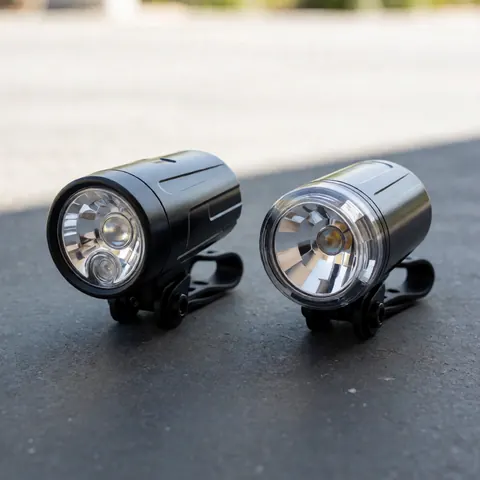 Deux phares de vélo noirs avec des lentilles claires, fixés sur des supports, posés sur une surface en béton gris, éclairés par une lumière naturelle neutre.