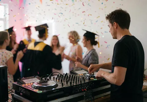 Un DJ professionnel mixe de la musique pendant une fête de fin d'études avec des gens qui dansent en arrière-plan, du confetti et des lumières vives.