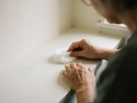 Une main adulte saisit un tissu en coton blanc sur une surface propre, éclairée par la lumière naturelle dans un studio aux murs blancs.