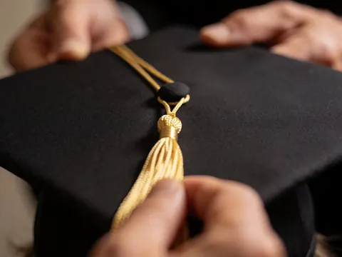 Détail de la frange dorée attachée à une casquette de graduation, mains ajustant le nœud sur fond flou avec éclairage chaleureux.