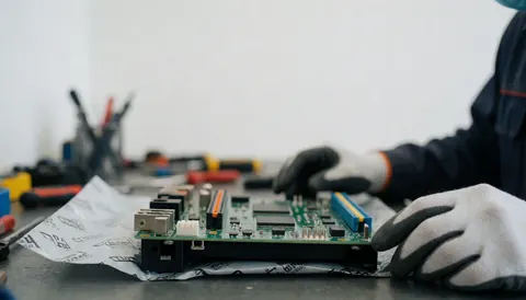 Technicien portant des gants de protection répare la carte de commande d'un lave-linge sur une table de travail dans un atelier avec des outils en arrière-plan flou.