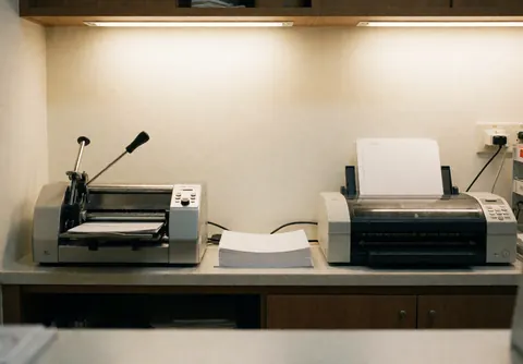 Poste de travail professionnel avec machine à relier et plastifieuse, pile de papier blanc, murs crème et éclairage chaleureux.