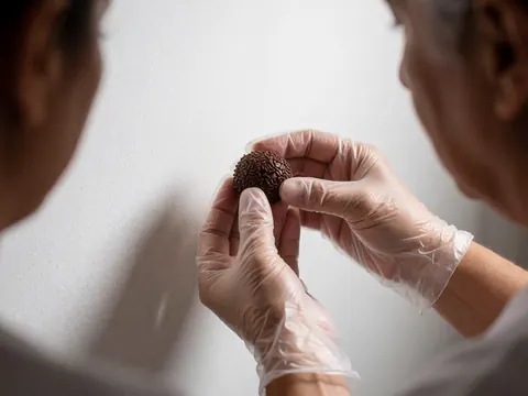 Mains portant des gants posant un petit gâteau au chocolat recouvert de paillettes colorées sur un fond blanc, vue rapprochée des détails.