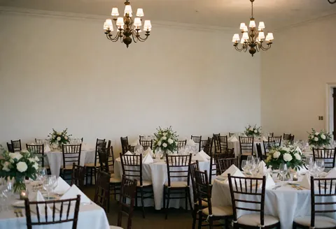 Salle d'événements pour mariage avec tables couvertes de nappes blanches, chaises en bois, centres de fleurs blanches et lustres illuminant l'espace vide et élégant.