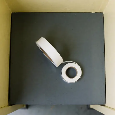 Rouleau de ruban PTFE blanc mat posé sur une table de travail grise, offrant un fond propre et une lumière douce pour la réparation d'appareils.