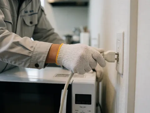 Un technicien portant un gant branche le câble d'un micro-ondes sur une prise dans une cuisine moderne au fond flou.