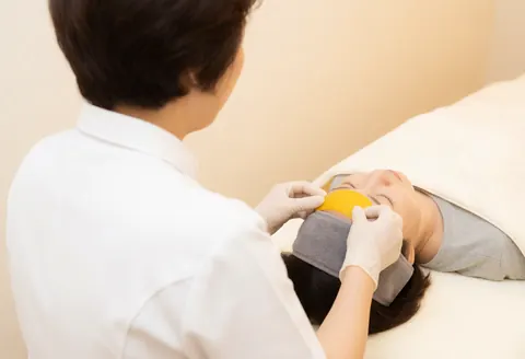 Une thérapeute en gants applique délicatement une tranche de fruit jaune sur le front d'une cliente les yeux fermés, dans un salon aux murs beige et à la lumière chaude.