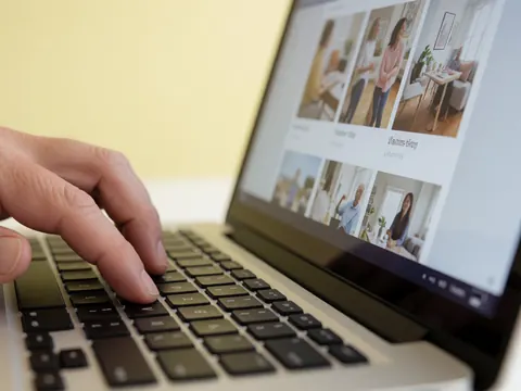 Une main adulte tape sur le clavier d'un ordinateur portable affichant une galerie de photos, avec un fond mur jaune pâle et une lumière douce professionnelle.