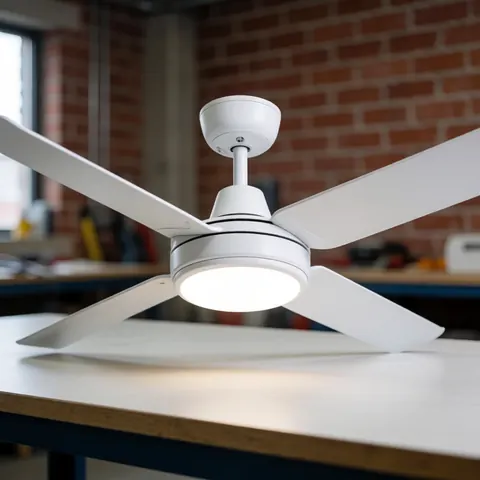 Ventilateur blanc moderne à quatre pales avec lumière LED intégrée posé sur un établi dans un atelier au mur de briques rouges.