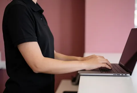 Vue latérale d'un bras et d'un clavier sur un ordinateur portable posé sur un comptoir, avec un mur rose en arrière-plan, évoquant un environnement de travail professionnel.