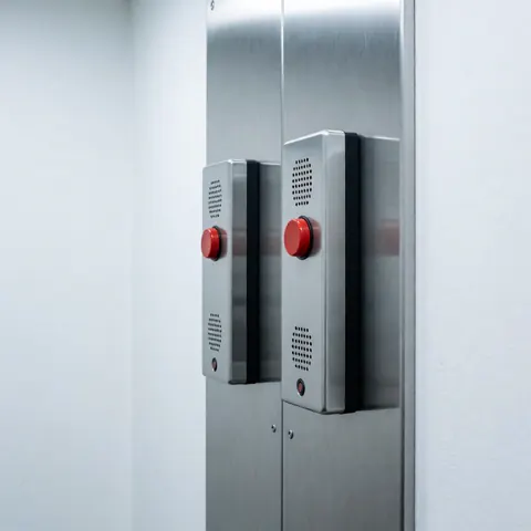 Deux interphones d'urgence avec boutons rouges fixés sur un panneau métallique dans une salle d'ascenseur. Éclairage froid, murs blancs et design industriel robuste.