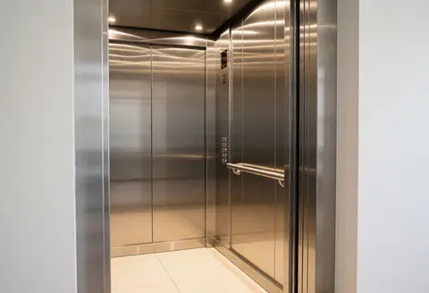 Intérieur d'un ascenseur moderne aux murs en acier inoxydable, avec une rambarde métallique et un sol carrelé blanc, éclairé par des LED chaudes, propre et sans personnes.