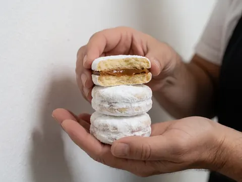 Des mains présentant une pile d'alfajores saupoudrés de sucre glace, l'un cassé révélant le remplissage crémeux au dulce de leche sur fond blanc.