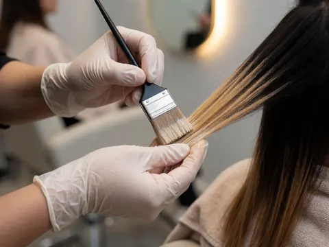 Des mains portant des gants appliquent de la teinture sur les cheveux avec un pinceau dans un salon de beauté à la lumière chaude et au fond flou.