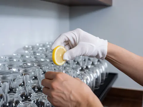 Une main portant un gant blanc dépose délicatement une tranche de citron fraîche sur le bord d'un verre. Plusieurs verres à vin sont alignés en arrière-plan sur un fond flou.