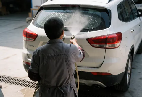 Un employé de lavage de voitures projette de l'eau sur la vitre arrière d'un SUV blanc sur un sol en béton avec des grilles de drainage.