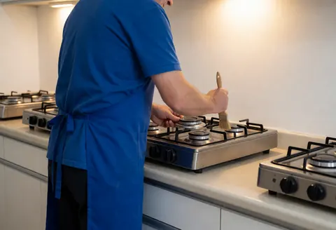 Un technicien en tenue bleue utilise une brosse pour nettoyer un brûleur de cuisinière dans une cuisine aux murs blancs et à l'éclairage chaleureux, dans un contexte professionnel et réaliste.