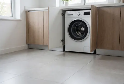 Lave-linge blanc à chargement frontal intégré dans un meuble de cuisine moderne avec sol carrelé gris et fenêtre laissant entrer la lumière naturelle.