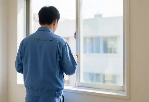 Un ouvrier en uniforme bleu vérifie le joint d'une fenêtre blanche installée dans un bureau aux murs clairs, éclairé par la lumière du jour.