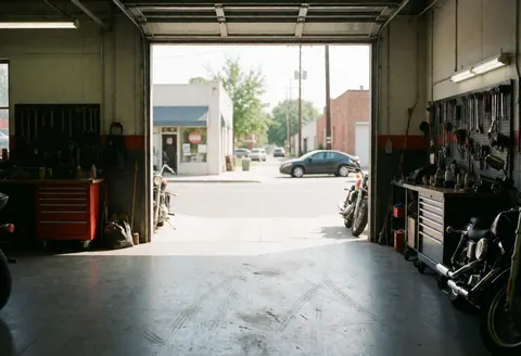 Façade et intérieur d'un garage de motos. Outils suspendus au mur, sol en béton marqué par des pneus, motos stationnées et rue avec voitures en arrière-plan.
