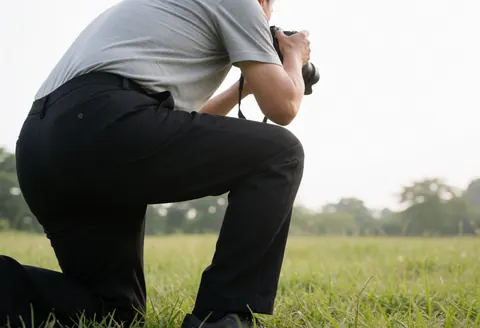Un photographe professionnel est agenouillé dans un champ verdoyant, capturant des images avec un appareil photo reflex. La lumière naturelle et douce éclaire la scène, illustrant un service de photographie en action.