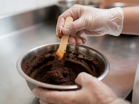 Une main portant un gant en nitrile mélange une pâte de café et de cacao dans un récipient métallique sur une table en acier inoxydable.