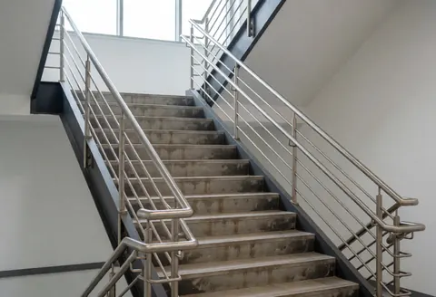 Escaliers de bureau avec garde-corps en métal et sols endommagés, prêts pour réparation et restauration esthétique, murs blancs et lumière naturelle.