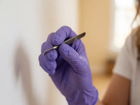 Gros plan d'une main portant un gant violet tenant un instrument de précision pour le soin des sourcils sur fond intérieur flou et chaleureux.