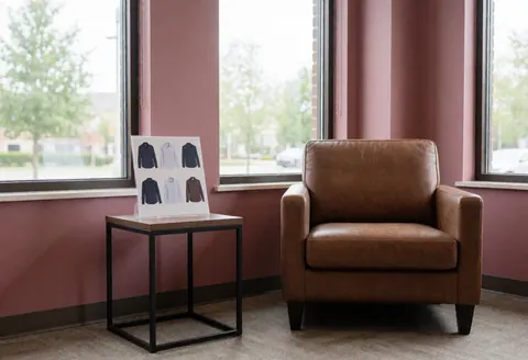 Salle d'attente avec un fauteuil en cuir marron, une table portant un catalogue de chemises, de grandes fenêtres laissant entrer la lumière naturelle et des murs couleur rose poudré.