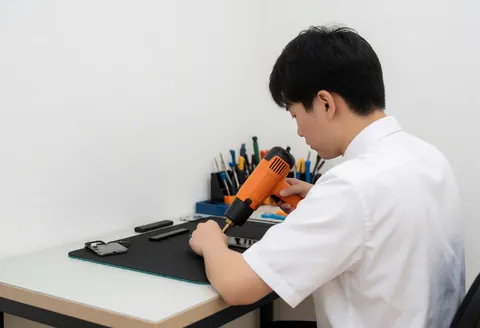 Un technicien utilise un pistolet à chaleur sur un écran de téléphone dans un atelier avec des outils et un fond blanc.