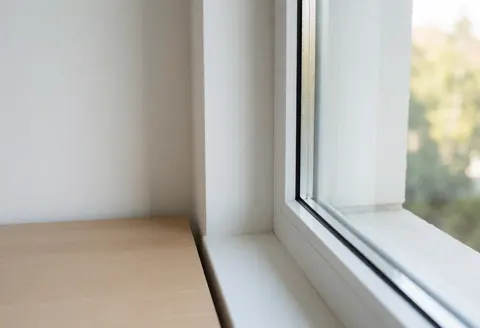 Vue d'une fenêtre d'office propre au cadre blanc, éclairée par la lumière du jour créant des reflets sur le verre clair et un bureau en bois naturel.